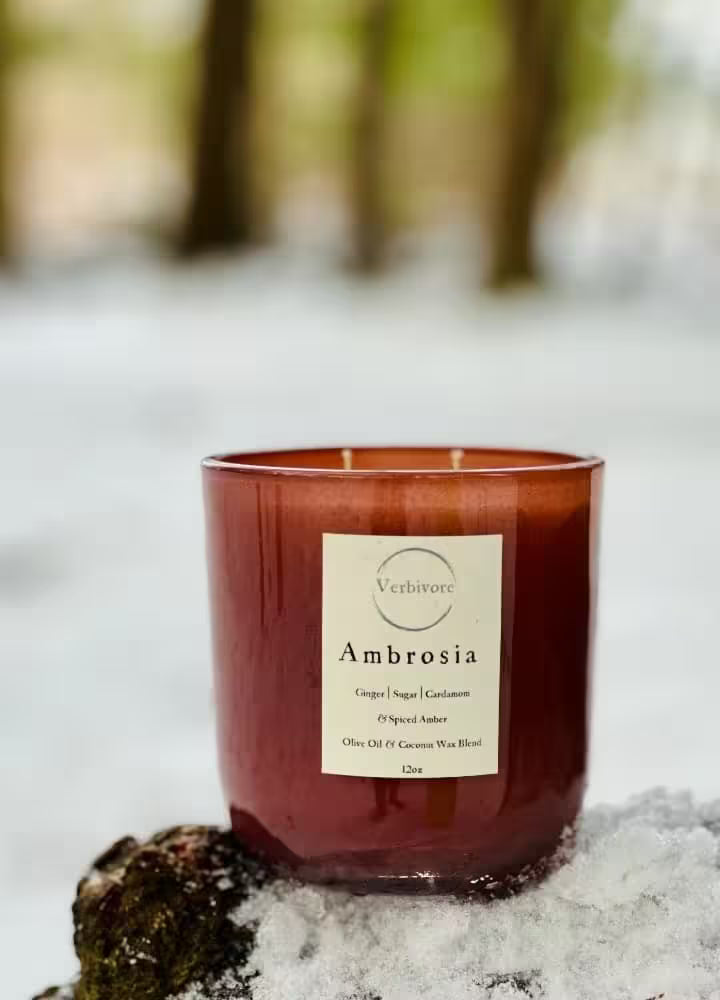 Red candle labeled 'Ambrosia' on a snowy surface with blurred trees in the background